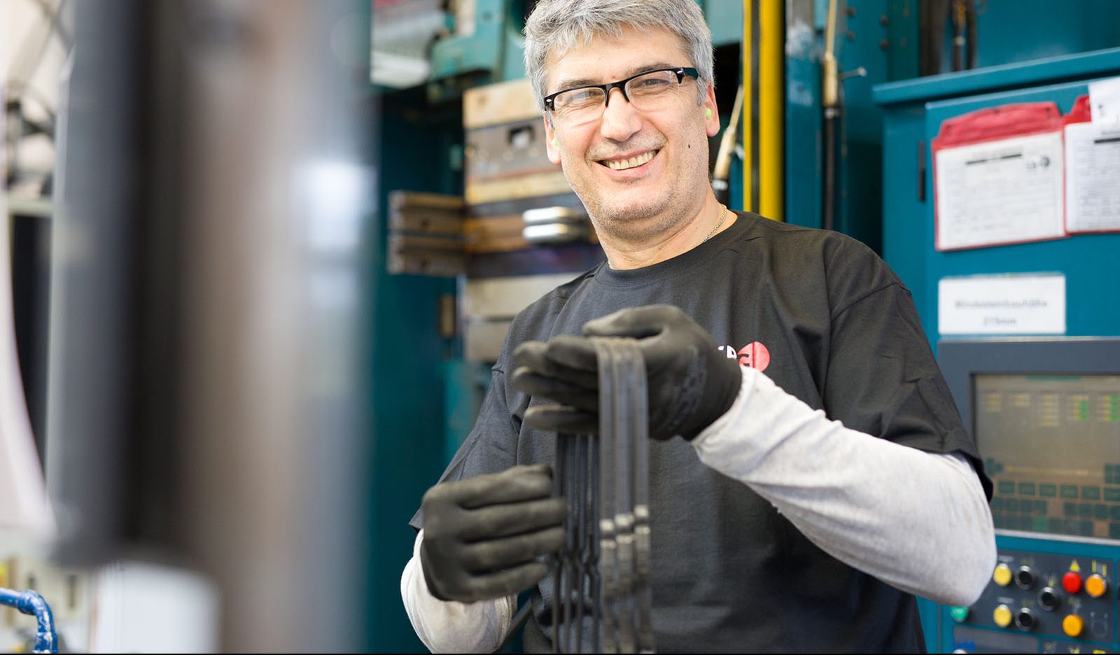 Mitarbeiter lächelt freundlich in die Kamera, während er in der Produktion gefertigte Gummi- bzw. Elastomerteile prüft